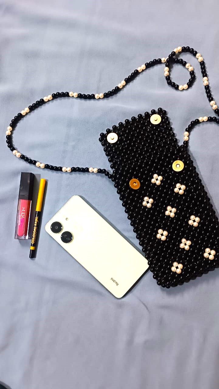 Handmade beaded black crossbody pouch with white floral bead design, holding a phone, placed on a blue cloth background next to lipstick and eyeliner.